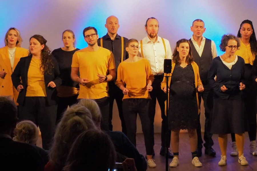 Ein Chor aus mehreren Männern und Frauen singt auf einer Bühne, gekleidet in gelb-schwarzer Kleidung. Die Sängerinnen und Sänger stehen in einer Reihe, teilweise mit expressiven Gesichtsausdrücken und Handgesten. Im Vordergrund ist das Publikum zu sehen.