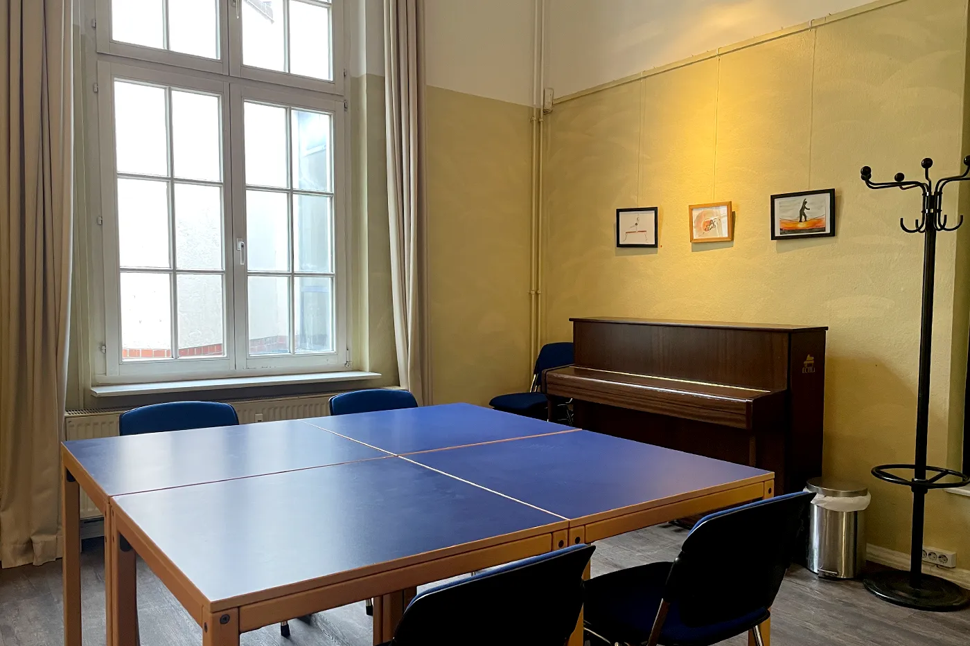 Offener Treff Ansicht mit großem blauen Tisch und schwarzem Klavier, durch ein großes Sprossenfenster fällt Licht in den Raum