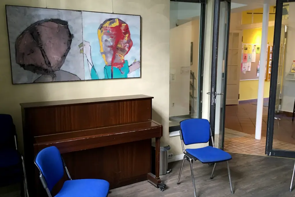 Offener Treff Ansicht mit zwei blauen Stühlen und schwarzem Klavier, bunte Bilder hängen an der Wand, eine Glastür führt nach draußen