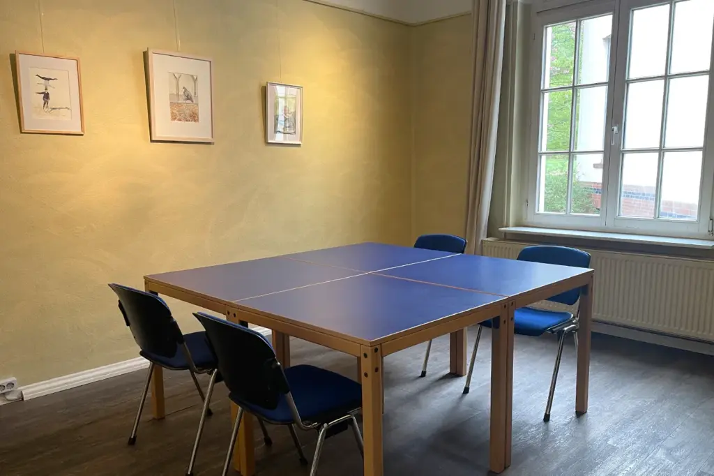 Offener Treff Ansicht mit großem blauen Tisch und gelber Wand, durch ein großes Sprossenfenster fällt Licht in den Raum
