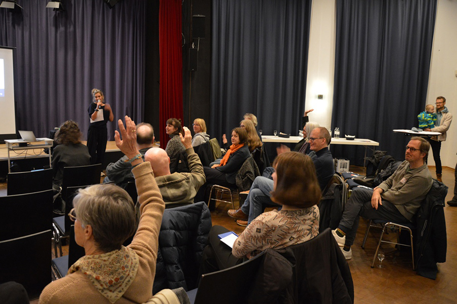 Eine Gruppe von Menschen sitzt in einem Saal und nimmt an einer Veranstaltung teil. Mehrere Personen heben die Hand, während eine Frau vorne mit einem Mikrofon spricht. Die Atmosphäre wirkt konzentriert und engagiert.