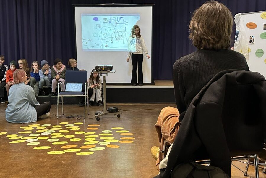 Ein Mödchen steht auf einer Bühne vor einer Leinwand, auf die eine Skizze projiziert wird, mehrere Kinder sitzen auf der Bühnenkante, zwei Erwachsene beobachten vom Zuschauerraum aus.