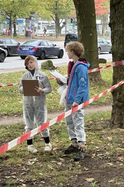 Zwei Jungen mit Klemmbrettern stehen in einem mit Flatterband abgesperrten Bereich einer Grünanlage am Rand einer mehrspurigen Straße.