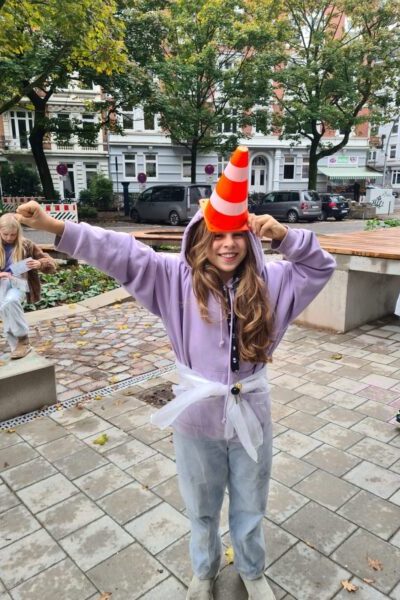 Ein Mädchen posiert lachend auf einem gepflasterten Platz mit Sitzbänken und Bäumen im Hintergrund und hält sich einen orange-weißen Leitkegel wie einen Zylinderhut auf den Kopf.