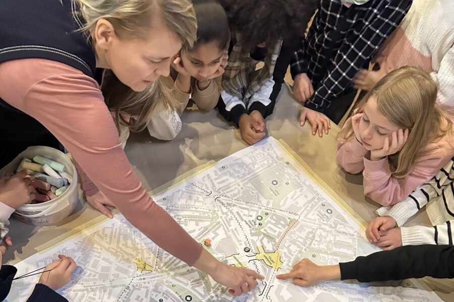 Eine Frau und mehrere Kinder beugen sich über einen großen Stadtplan, der mit Pauspapier überklebt ist. Areale sind durchgepaust, die Erwachsene und ein Kind zeigen mit den Fingern darauf.