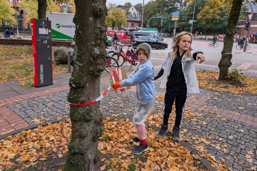Kind mit transparentem Maler-Schutzanzug über der Kleidung bindet auf ienem innerstädtischen Platz ein rot-weißes Absperrband um einen Baum, daneben steht ein Mädchen und zeigt nach vorne.