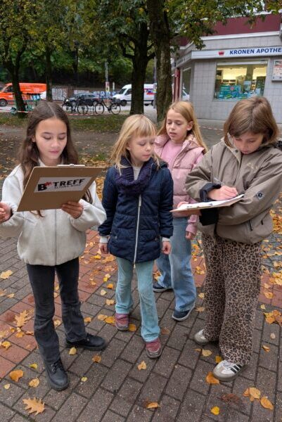 Vier Mädchen stehen auf einem gepflasterten Gehweg, der mit Herbstlaub bedeckt ist. Zwei der Kinder halten Klemmbretter in den Händen, eines davon trägt die Aufschrift 'B TREFF'. Im Hintergrund sind Bäume, Fahrräder und ein Gebäude mit der Aufschrift 'KRONEN APOTHEKE' zu sehen.