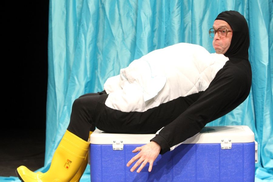 Ein als Pinguin verkleideter Schauspieler richtet sich auf einer Tiefkühltruhe aus dem Liegen auf