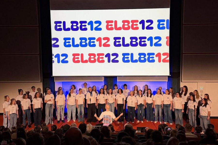 Der Chor singt auf einer Bühne, dahinter eine Leinwand mit dem sich mehrmals wiederholenden Schriftzug "Elbe12"