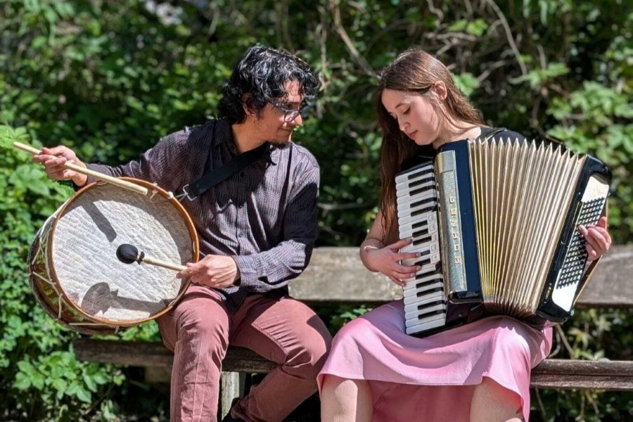 Das Duo sitzt auf einer Bank und musiziert an Trommel und Akkordeon
