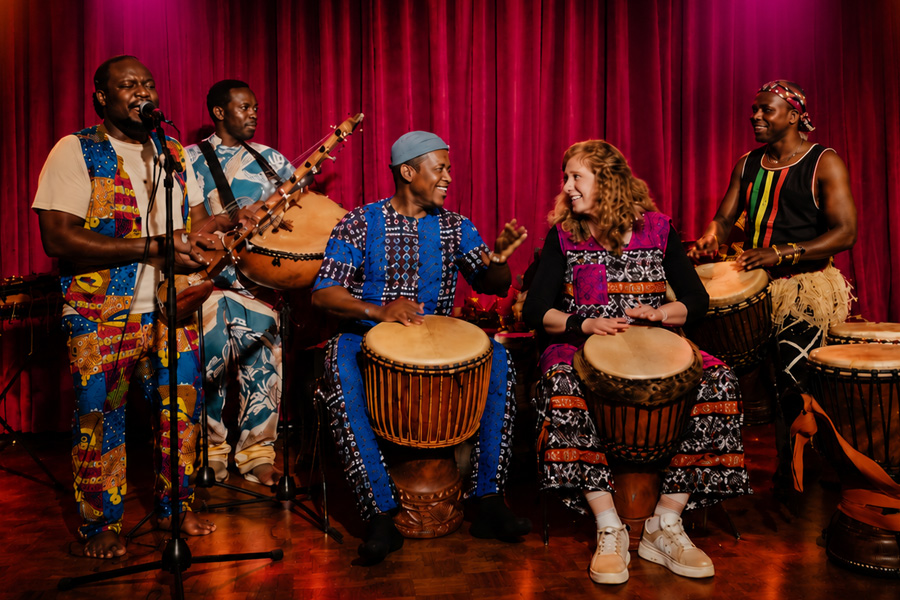 Billy Konate und Sylvia Franke trommeln gemeinsam mit drei Freunden vor einem roten Vorhang auf der Bühne